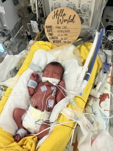 Newborn baby Max lying in a NICU incubator surrounded by medical equipment, wearing a tiny hat and oxygen support.